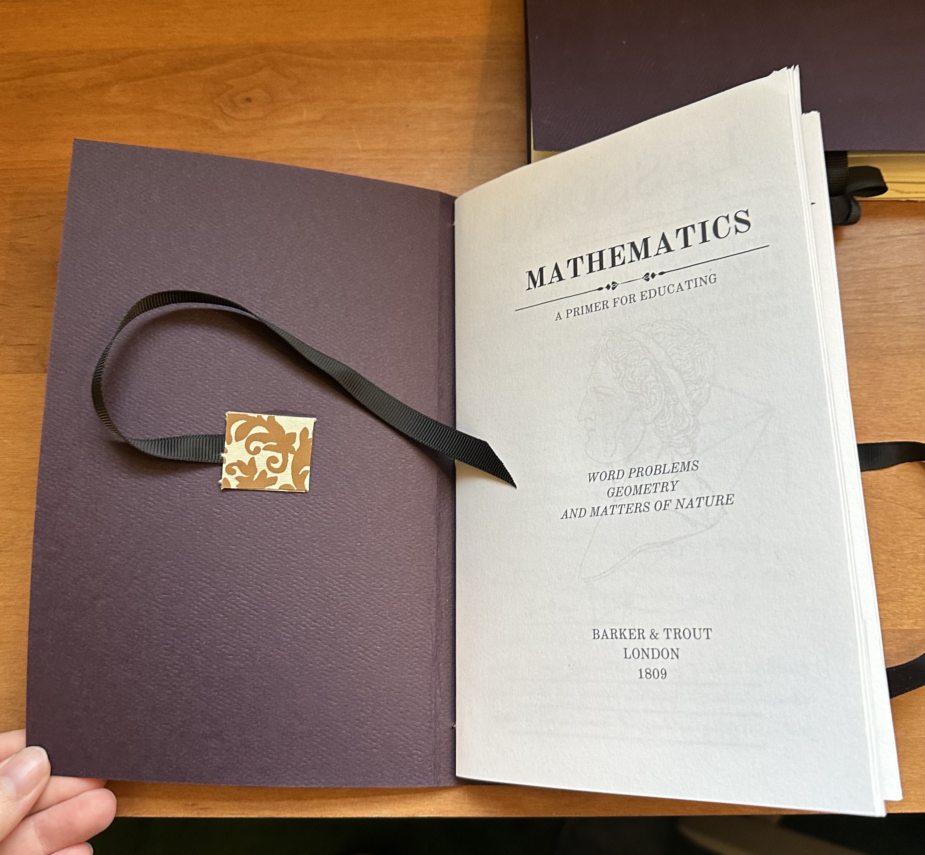 The study book lays open to the first page. On the back of the cover of the book is a square of decorative paper holding a ribbon down that stretches out to tie the book together. On the first page reads "MATHEMATICS" with other title-page looking stuff below but it's unreadable.