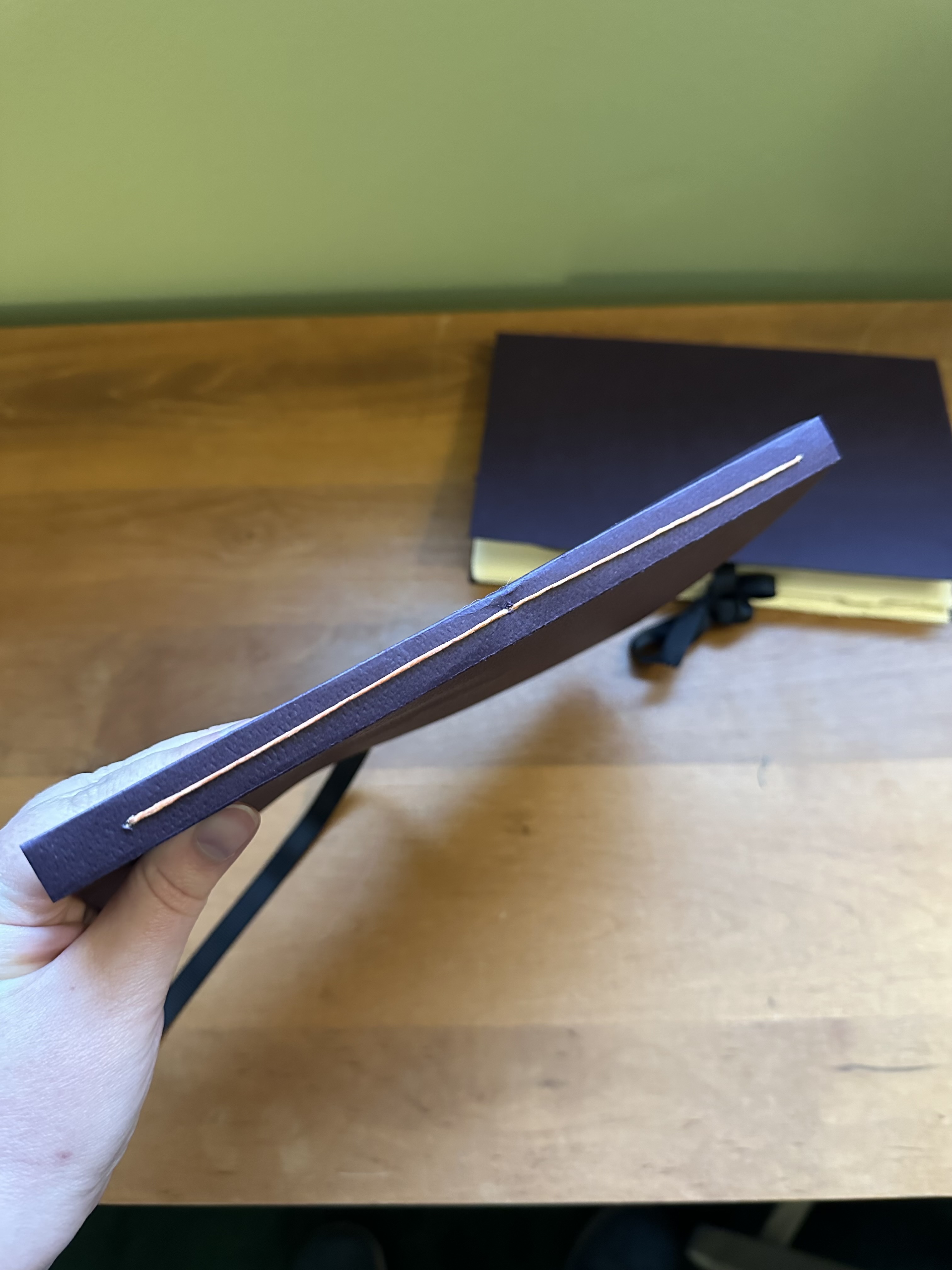 A hand holding up the spine of a purple book with orange thread holding it together. One that looks similar to it is on the table below.