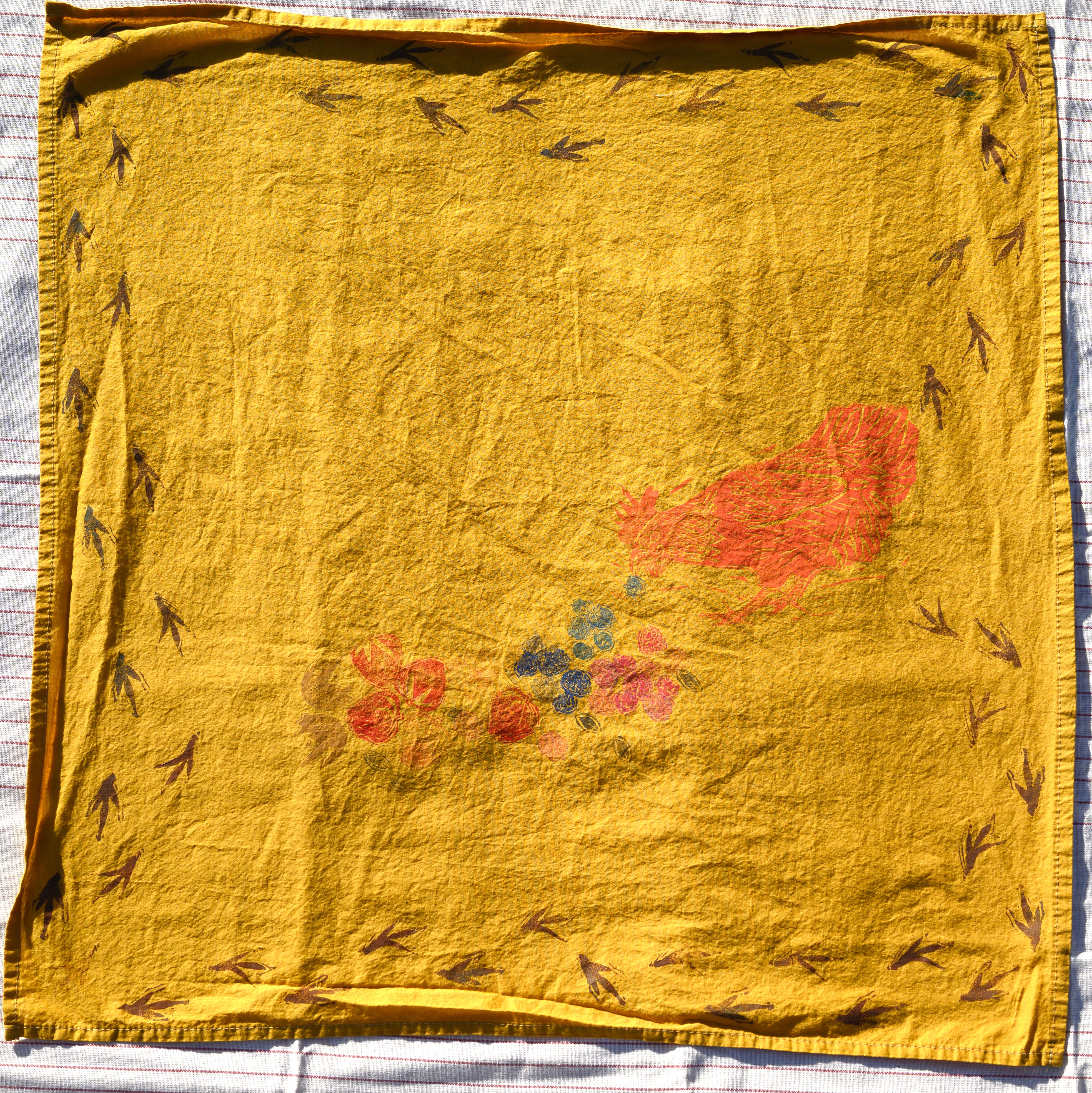 An overview of a mustard yellow bandana. There are borwn chicken prints as a border. There is a red-ish chicken near the center corner leaning down, tail in the air, to eat blueberries, raspberries, & strawberries.