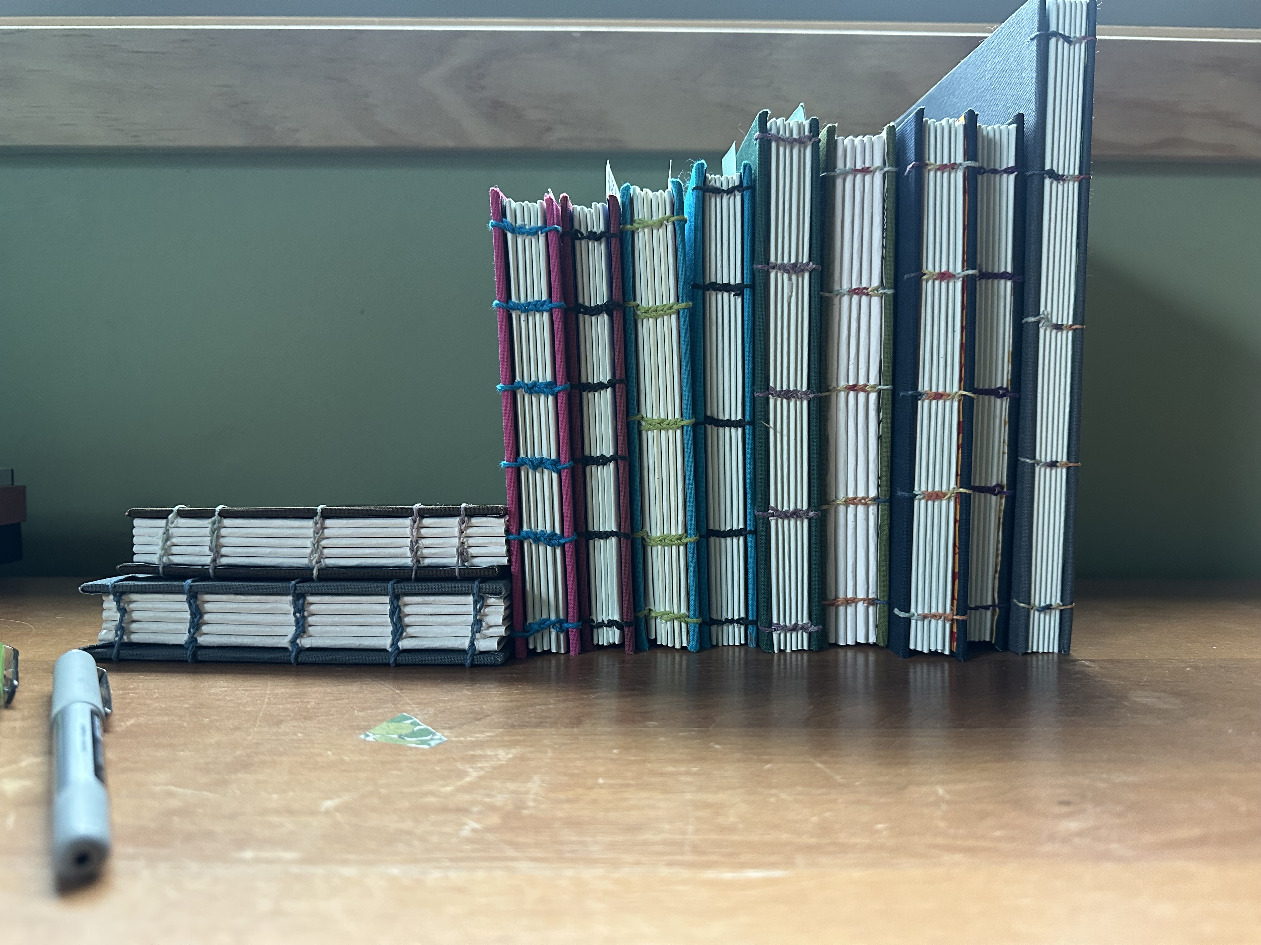 9 coptic bound hard cover books with various colors of covers sitting vertically. There are two sitting on their sides, laying horizontally one on top of the other on the left.