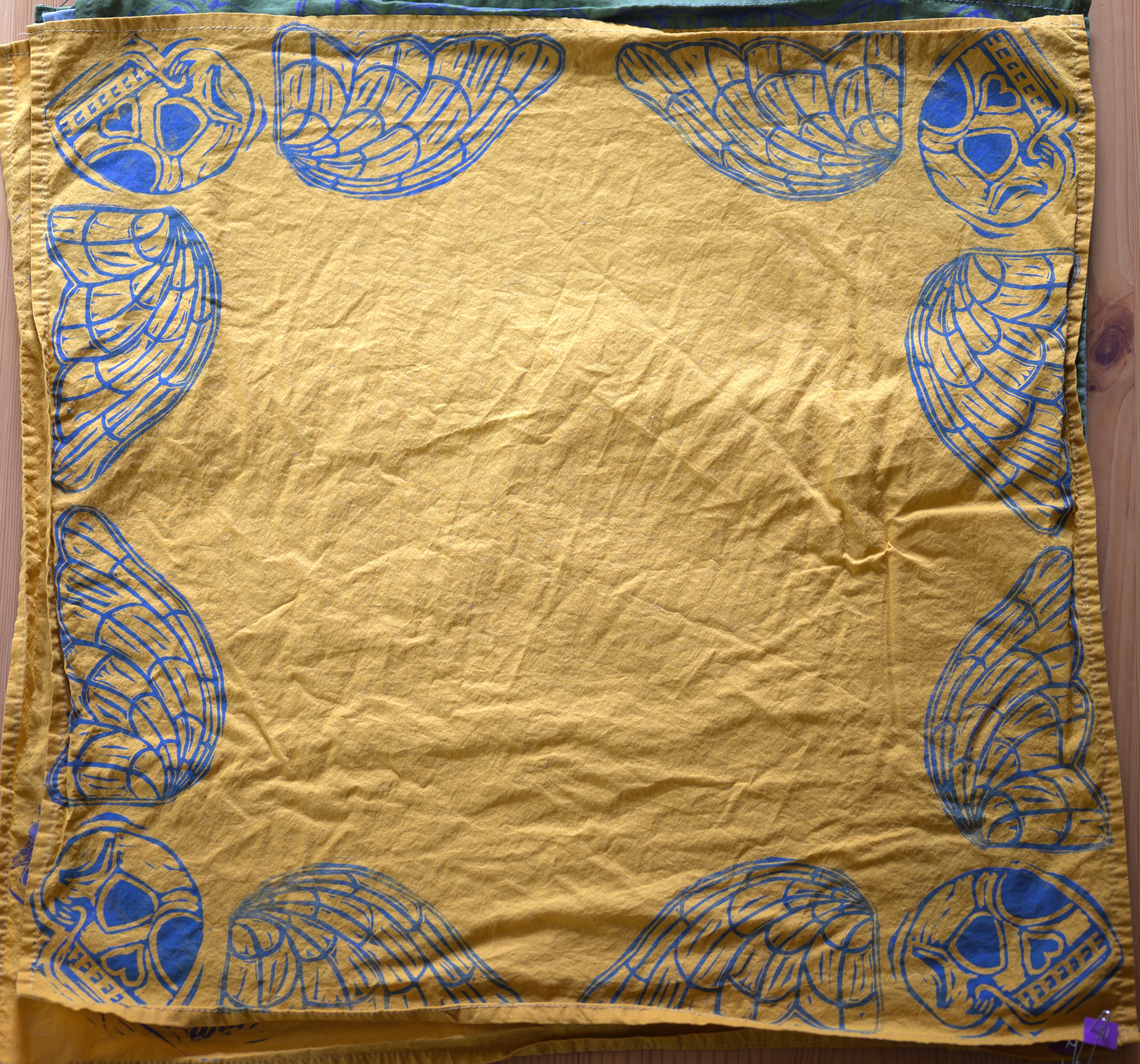 An overview of a mustard yellow bandana. There is a blue death's head in each corner (skull with angelic wings), taking up essentially the border of the bandana.