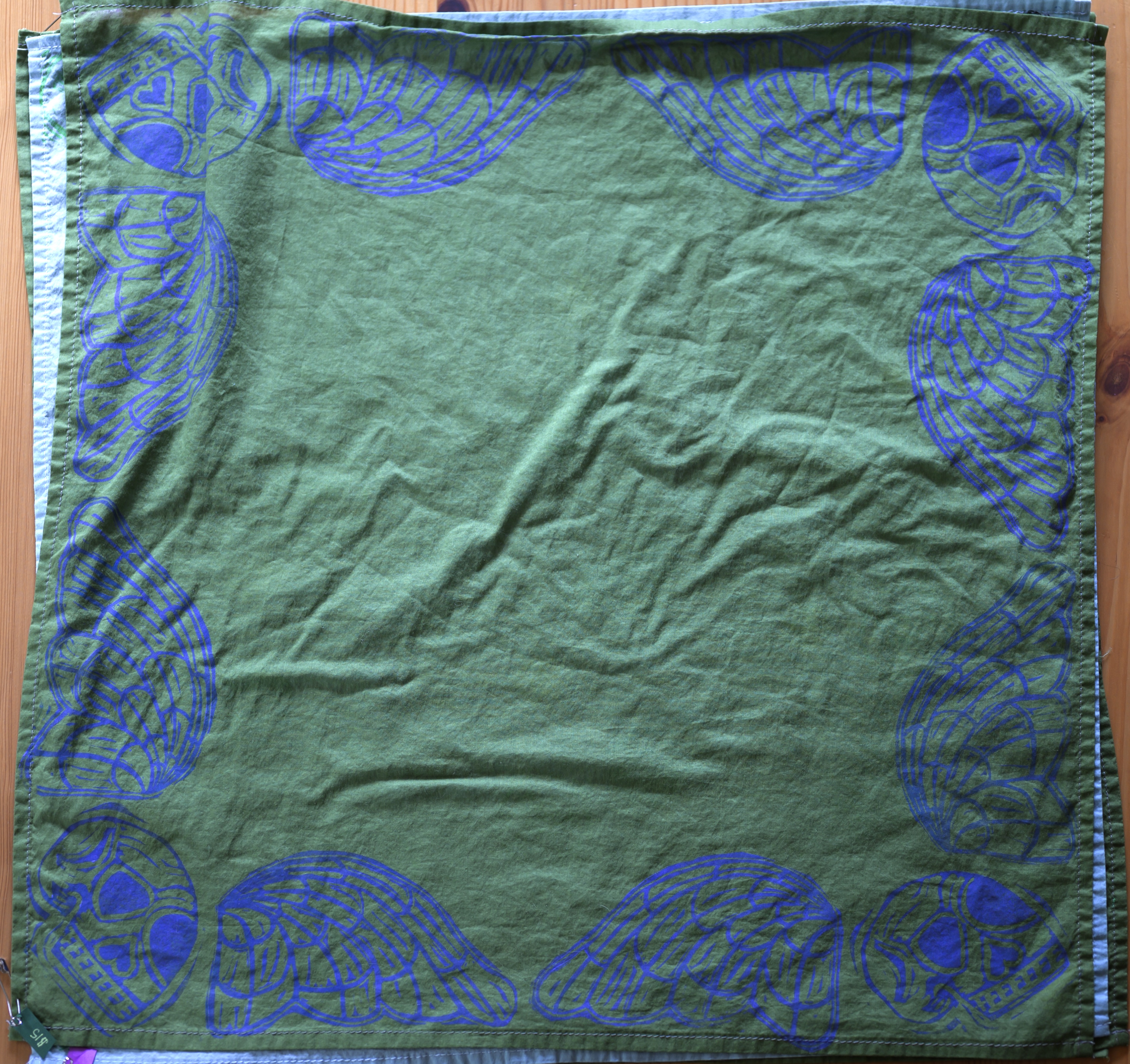 An overview of a deep green bandana. There is a purple-y blue death's head in each corner (skull with angelic wings), taking up essentially the border of the bandana.