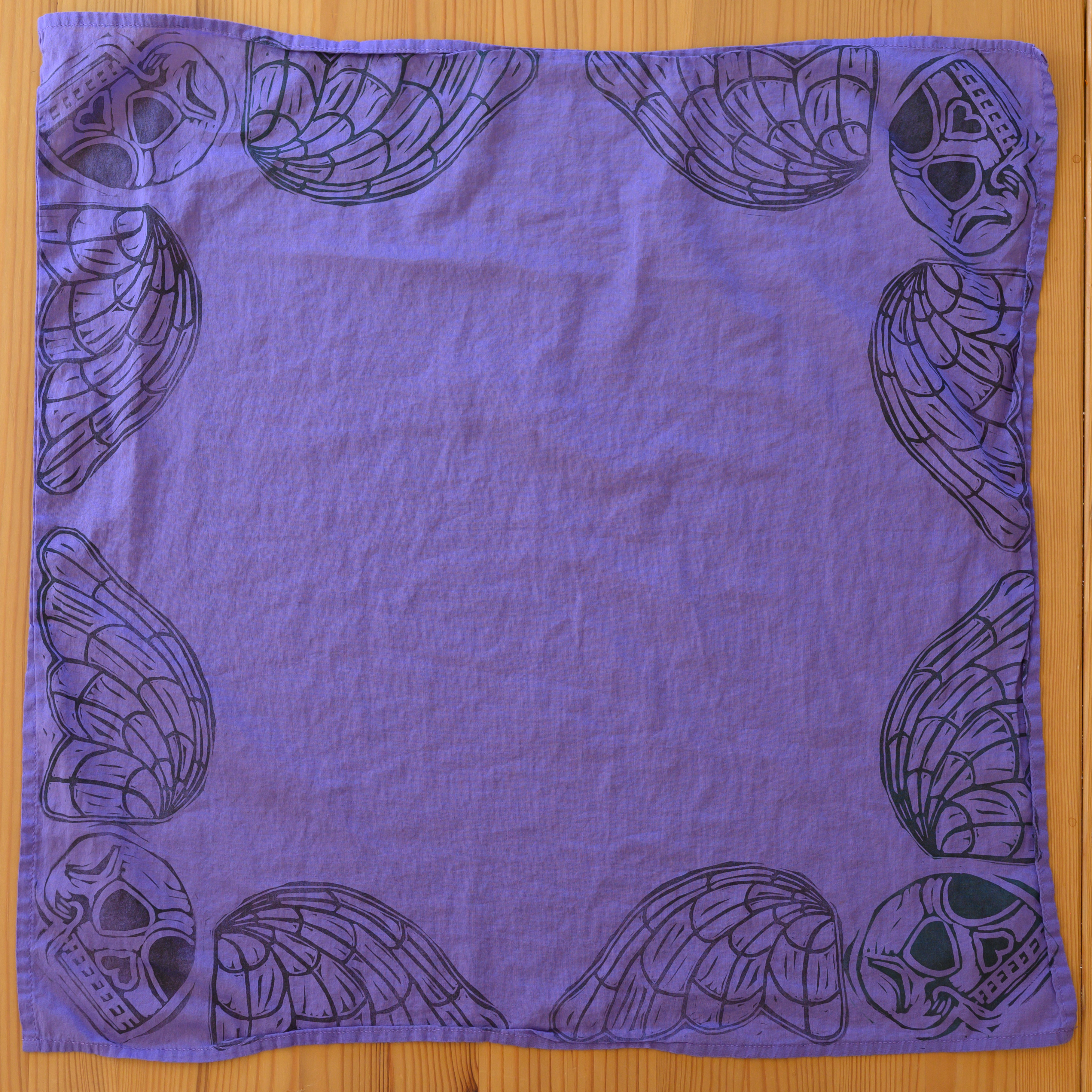 An overview of a purple bandana. There is a black death's head in each corner (skull with angelic wings), taking up essentially the border of the bandana.