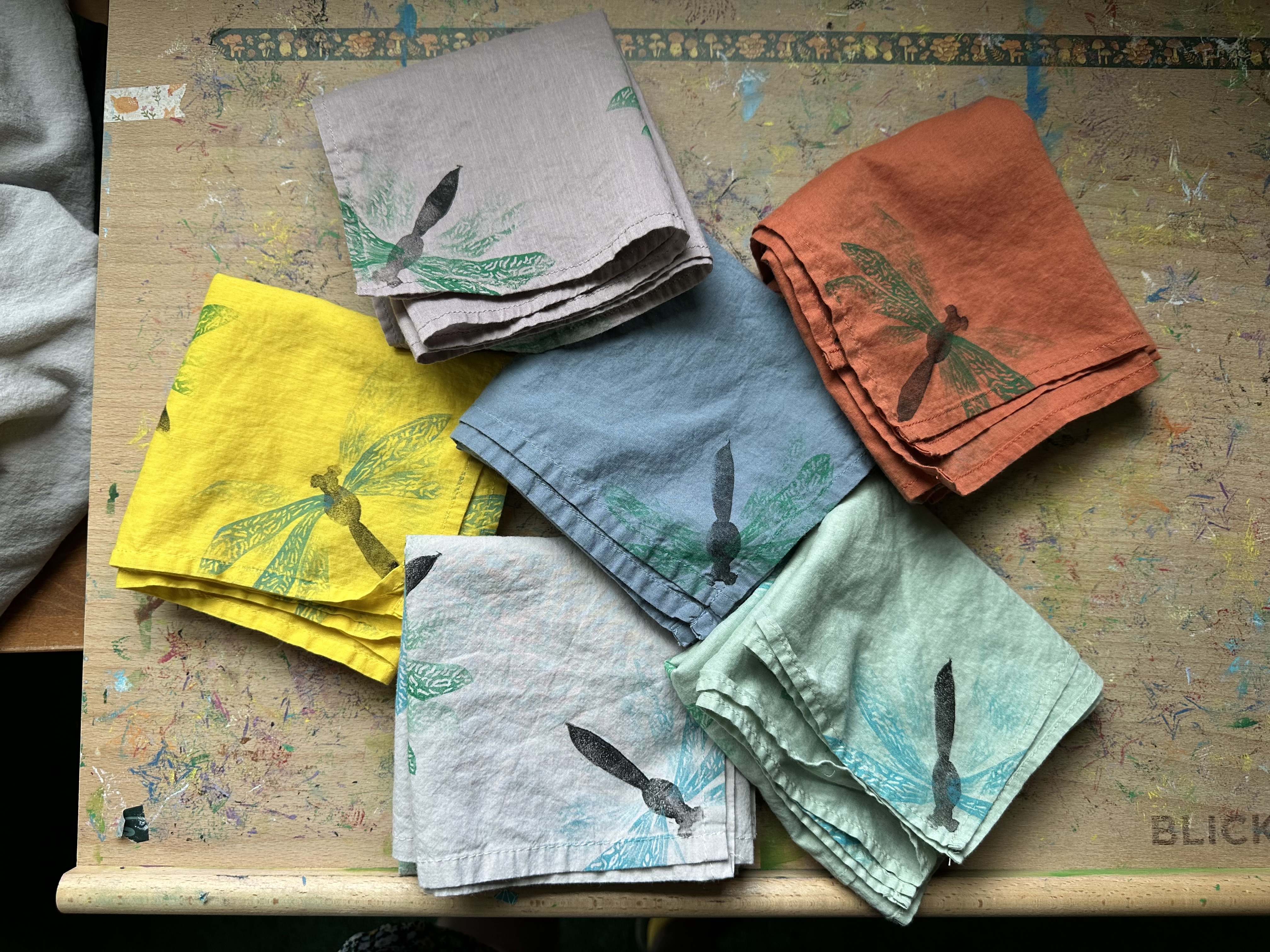 A look from above at 6 bandanas with dragonflies on each corner. Some are facing the corner, some are not, some have blue wings, some green. The bandanas are yellow, pale dusky pink, earthy orange, faded blue, light grey, and faded green.