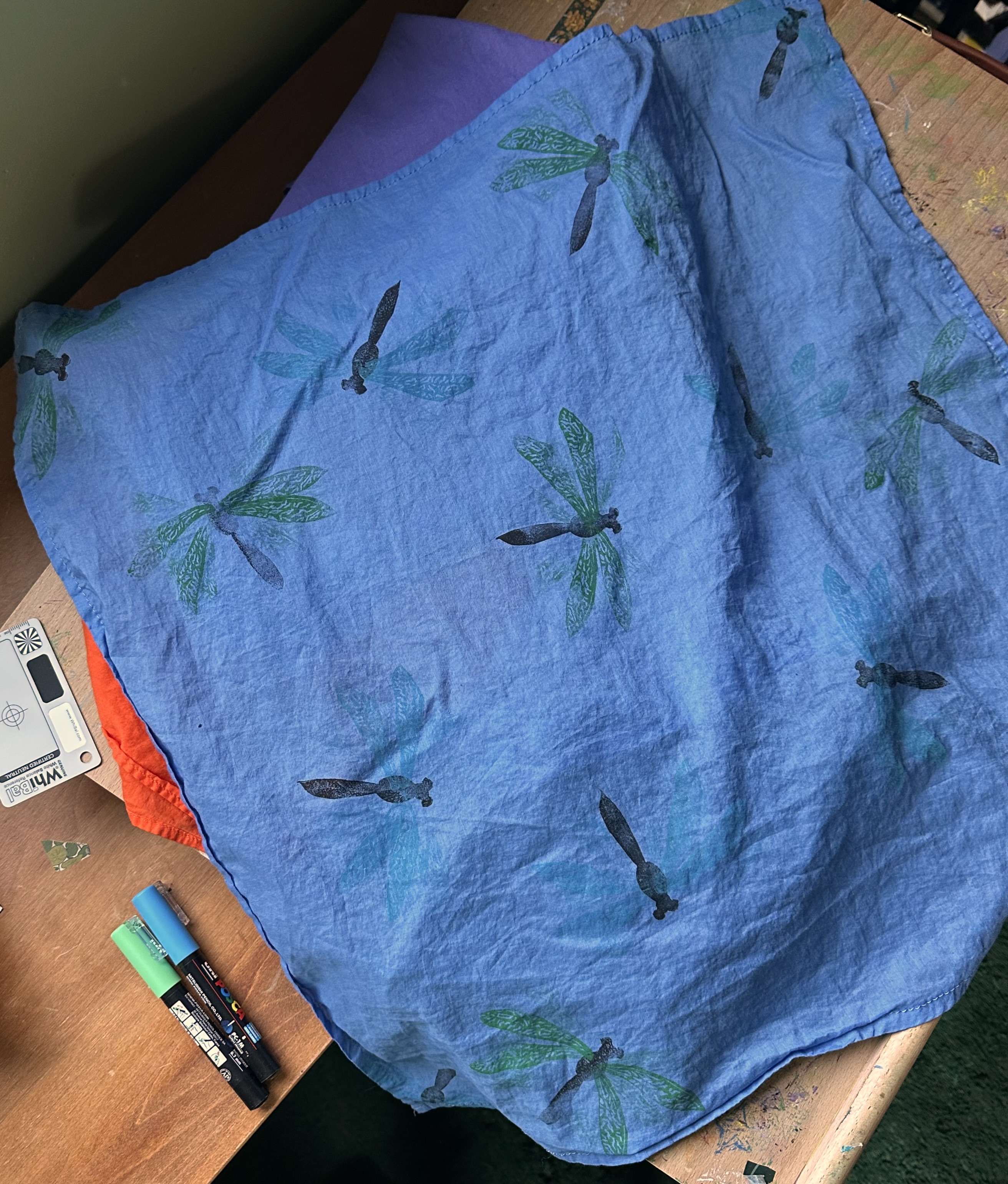 A close up of a bandana from above. It's a periwinkle blue with dragonflies pointing in different directions over it sporadically. Some have green wings, some blue.