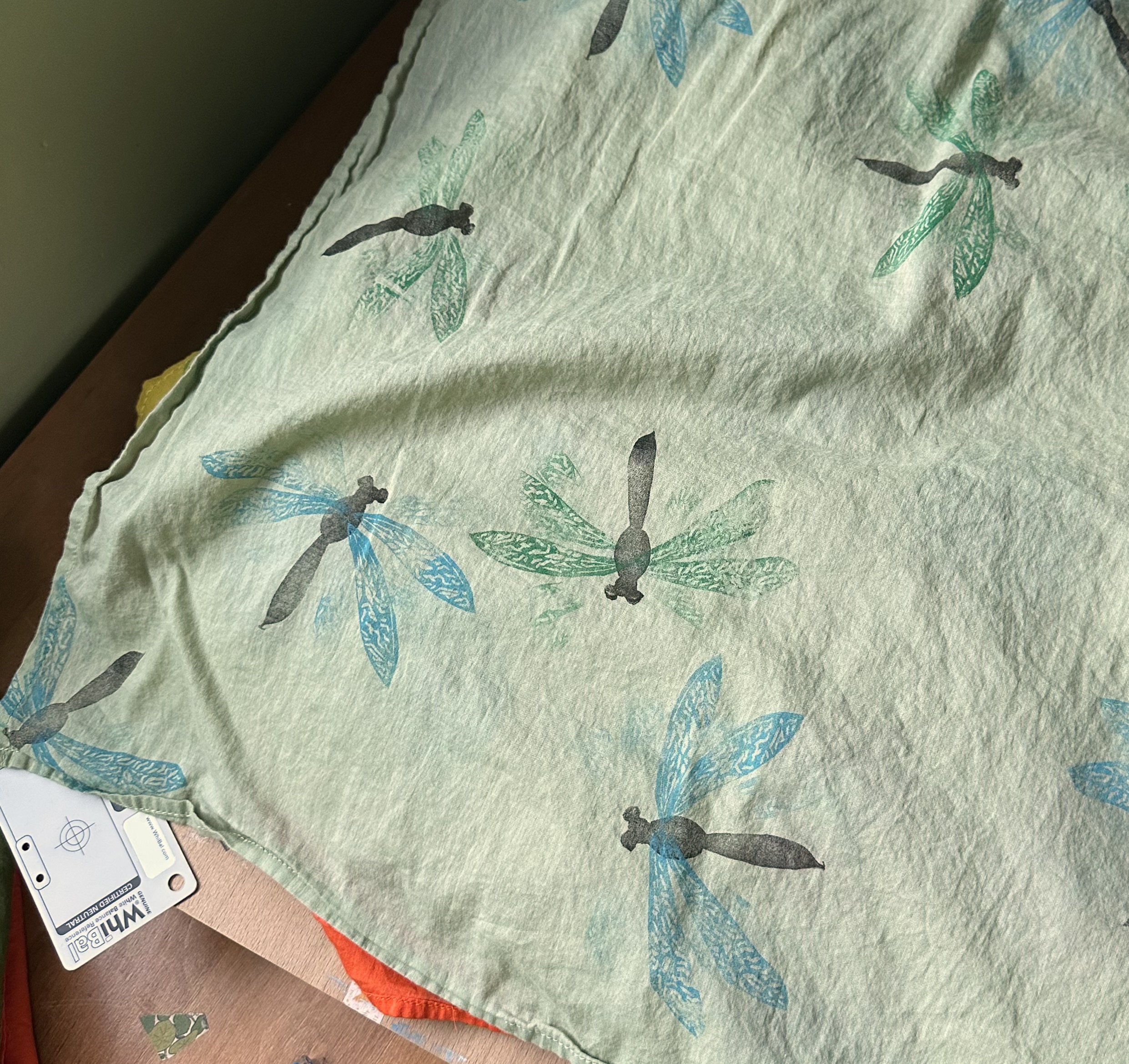 A close up of a bandana from above. It's dusky green with dragonflies pointing in different directions over it sporadically. Some have green wings, some blue.