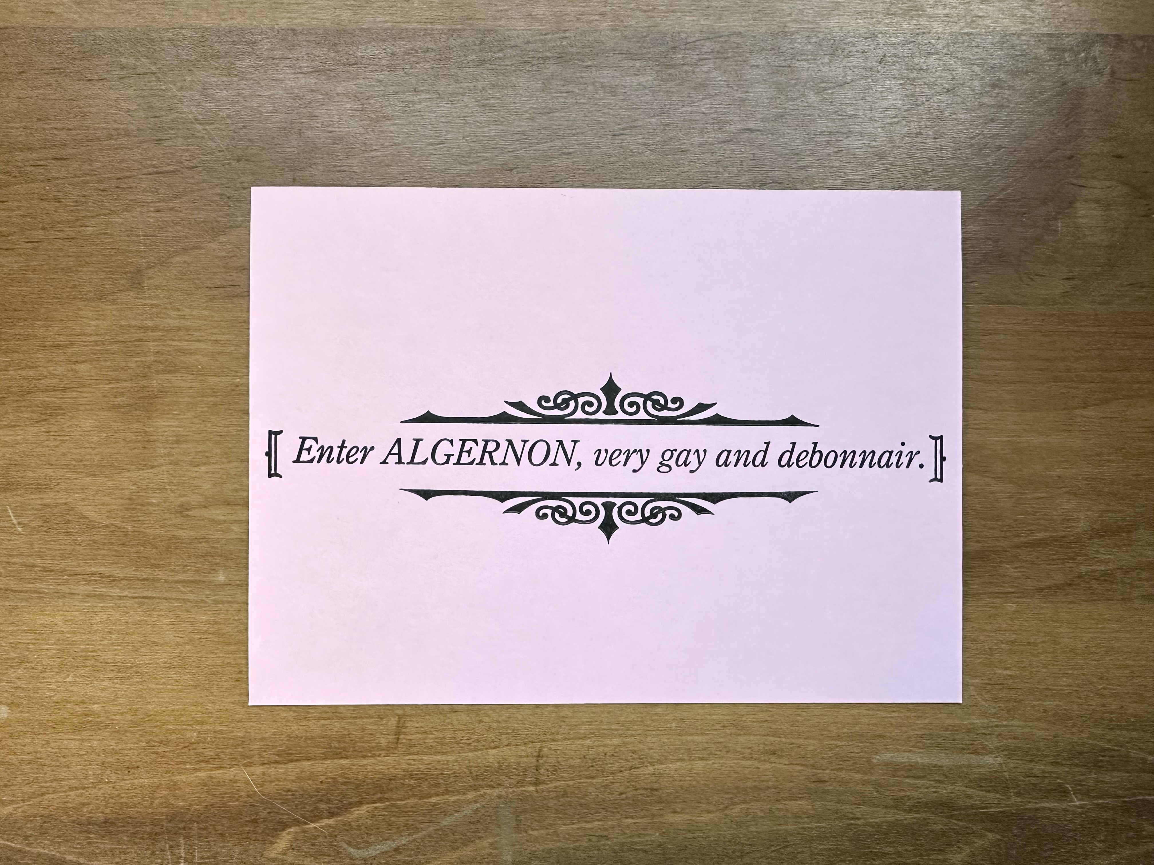 On a light pink rectangular piece of paper, [Enter ALGERNON, very gay and debonnair.] is printed in black in italics. Above and below are decorative swirls and lines.