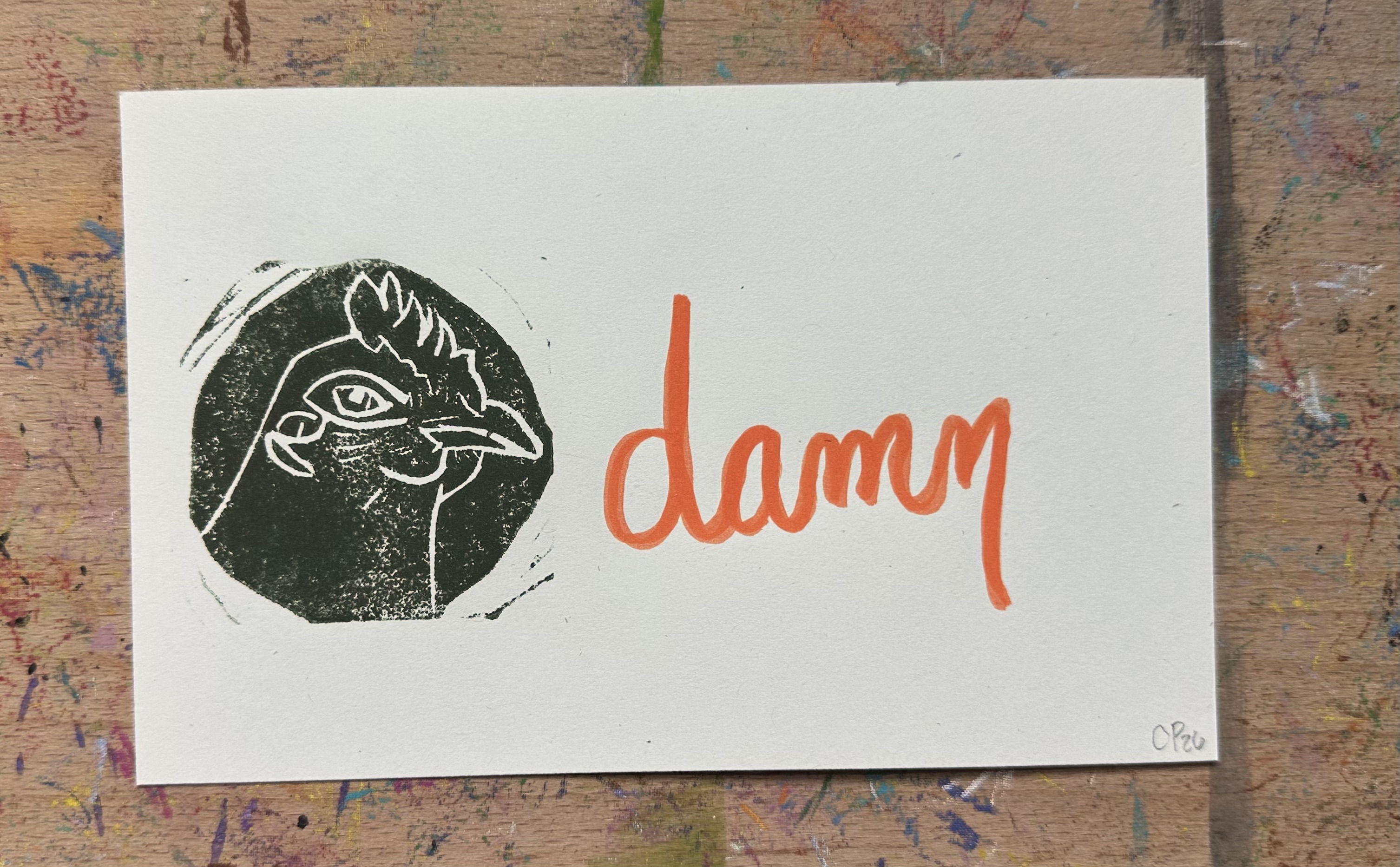 Rectangular piece of off white paper. A dark green/brown block print of a chicken in profile witha knowing look on the left. "Damn" written on the right.