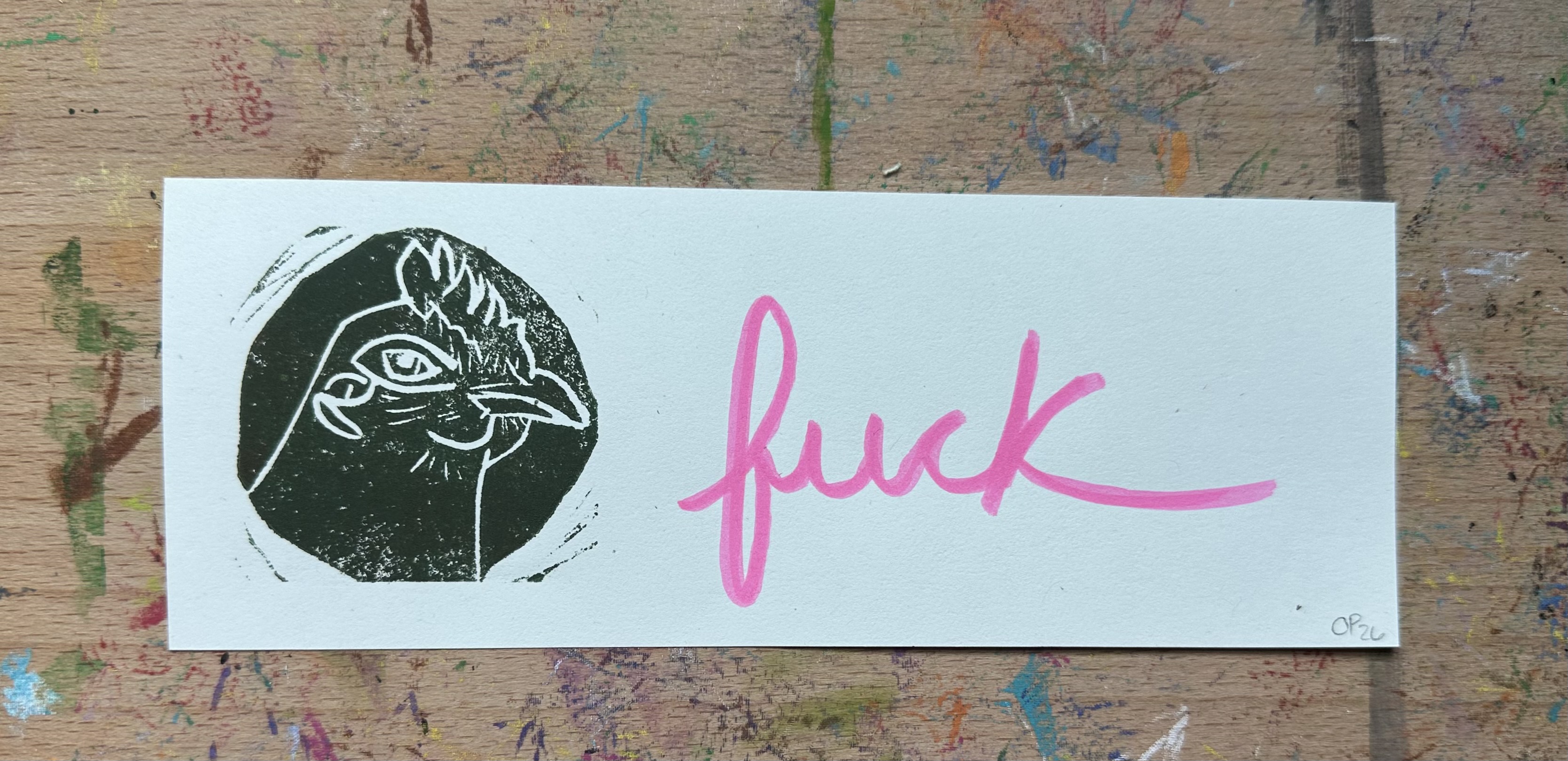 Rectangular piece of off white paper. A dark green/brown block print of a chicken in profile witha knowing look on the left. "fuck" written on the right.