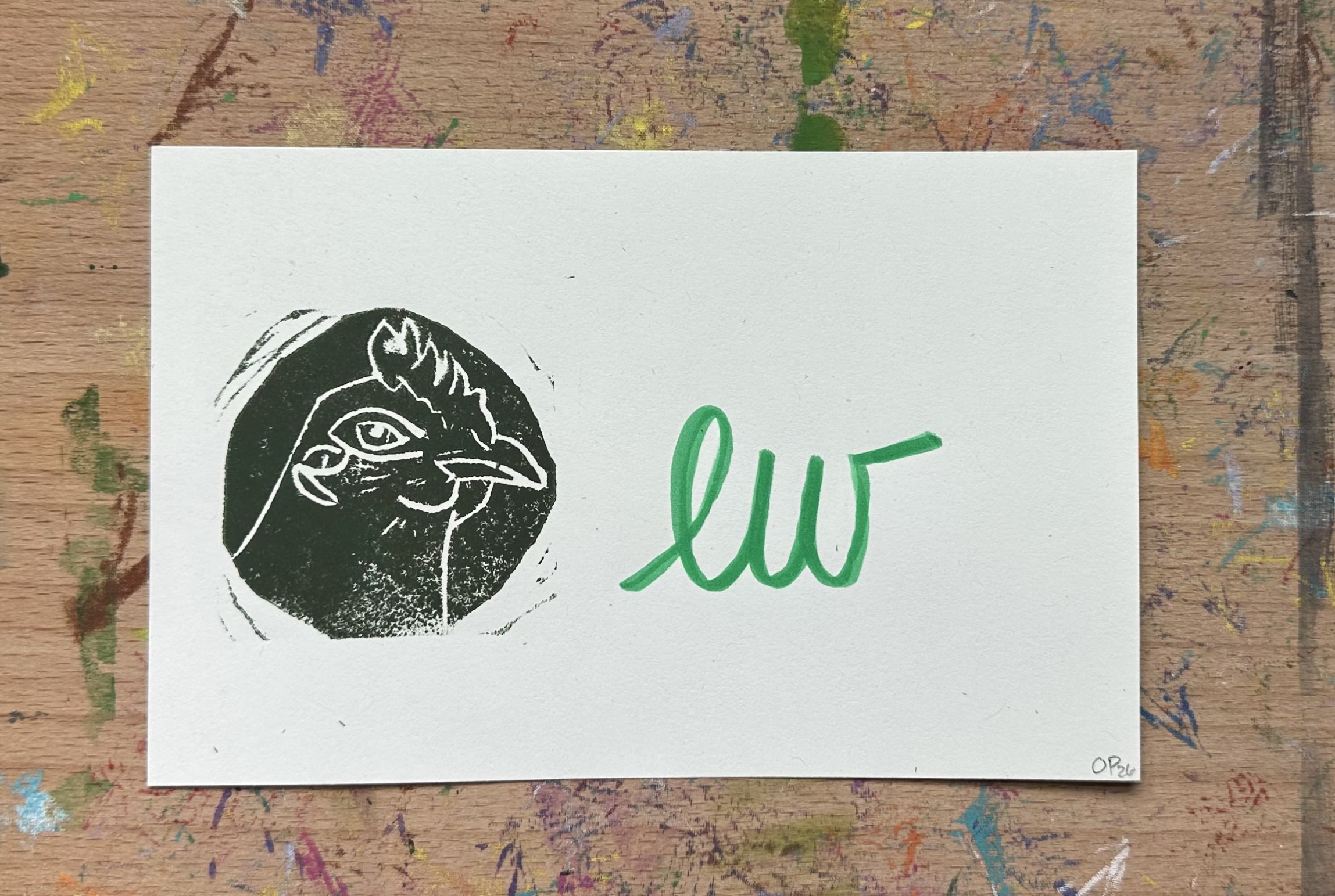 Rectangular piece of off white paper. A dark green/brown block print of a chicken in profile witha knowing look on the left. "ew" written on the right.