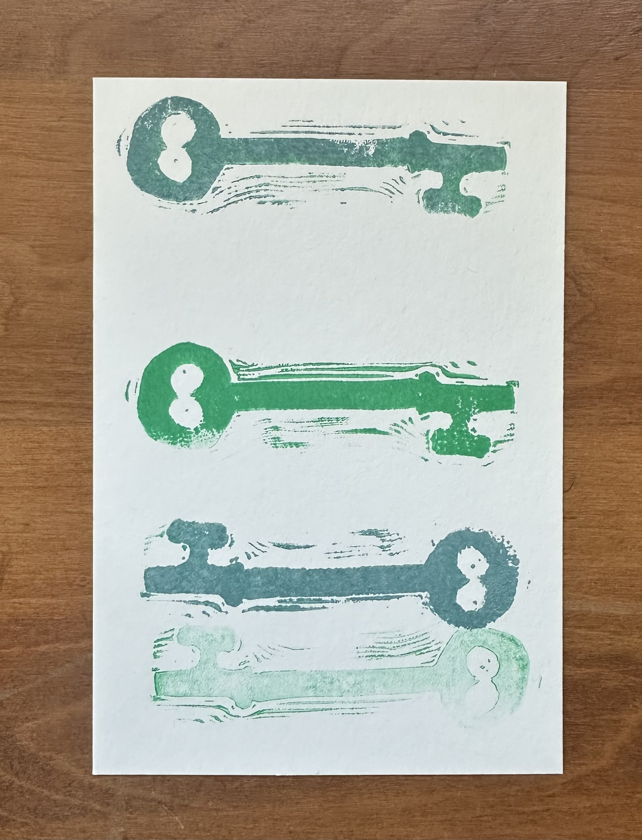 A white rectangle lays on a wood surface vertically. At the top is a block print of a skeleton key in a blue-ish green with the top of the key on the left. There's a noticible space below then another facing the same way but in bright green. A small space follows, then two more keys both with the top to the right. The first one is blue gray and the bottom one is green but only lightly so, like the printmaker didn't ink up the block again to print.