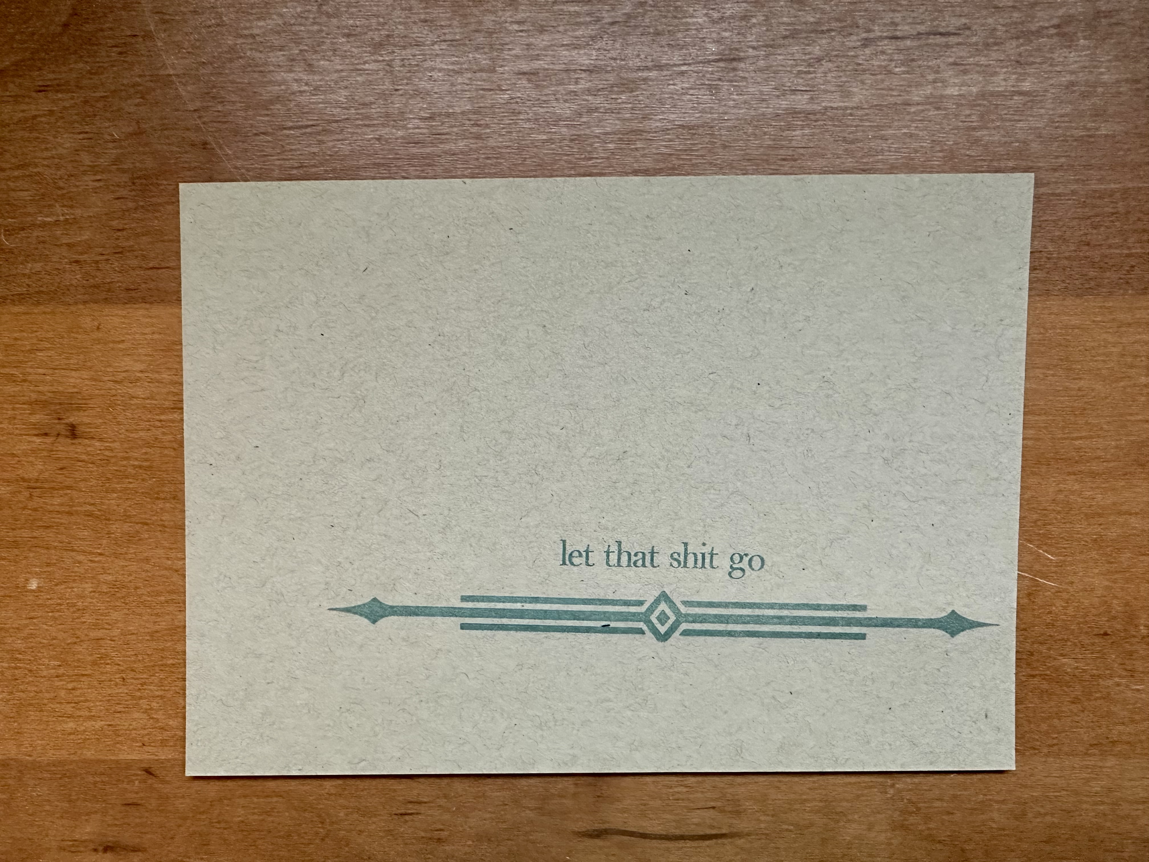On light brown paper, near the bottom it says "let that shit go" with an ornamental series of lines directly underneeth in green ink. The print does not line up with the paper.
