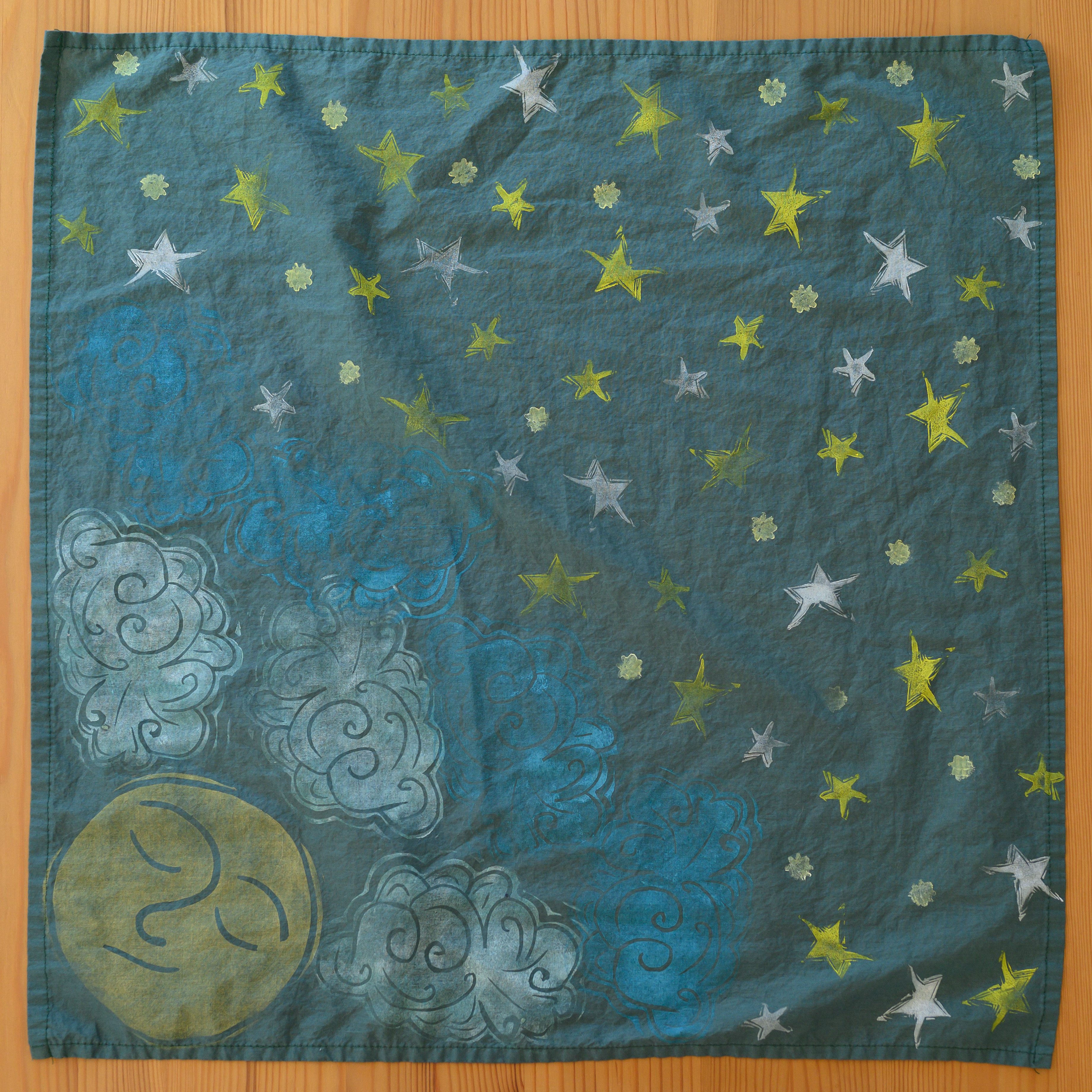 A close up of a hunter green bandana from above. Most of it is covered in yellow and white stars. In the left corner is a sleepy yellow moon face with white and blue clouds around it.