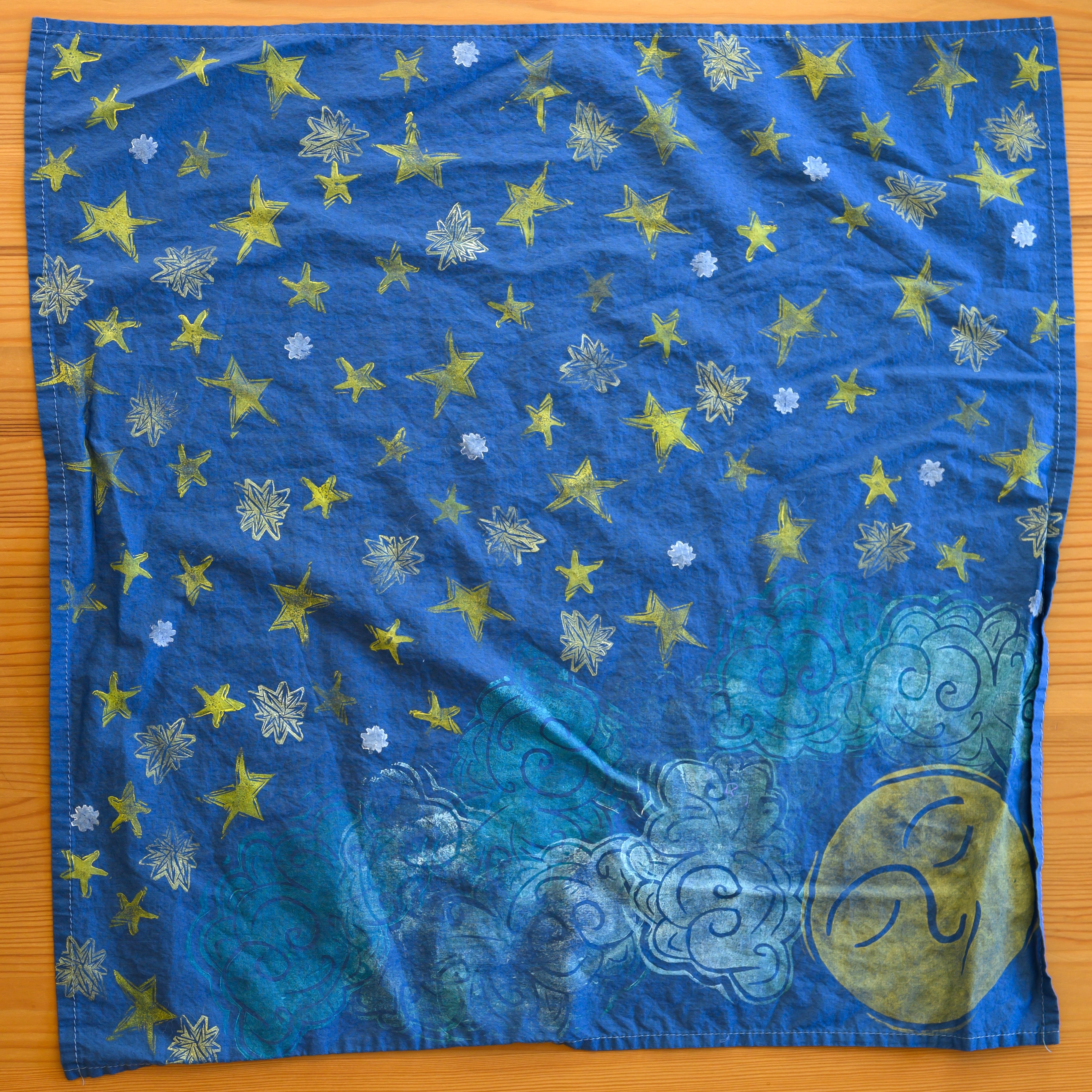 A close up of a navy bandana from above. Most of it is covered in yellow and white stars. In the right corner is a sleepy yellow moon face with white and blue clouds around it.