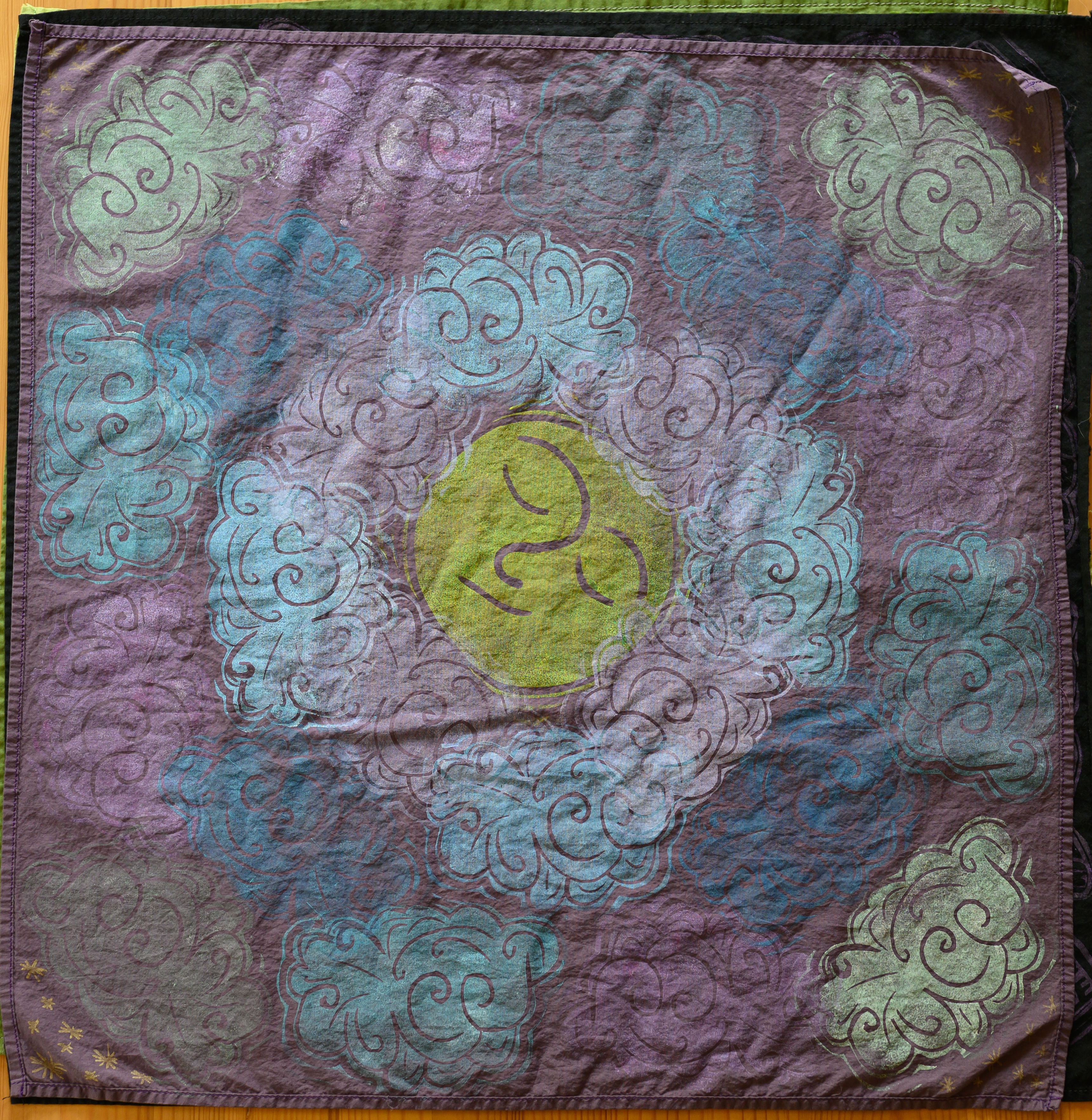 A close up of a purple bandana from above covered in clouds of various colors. In the center is a sleepy yellow moon face.