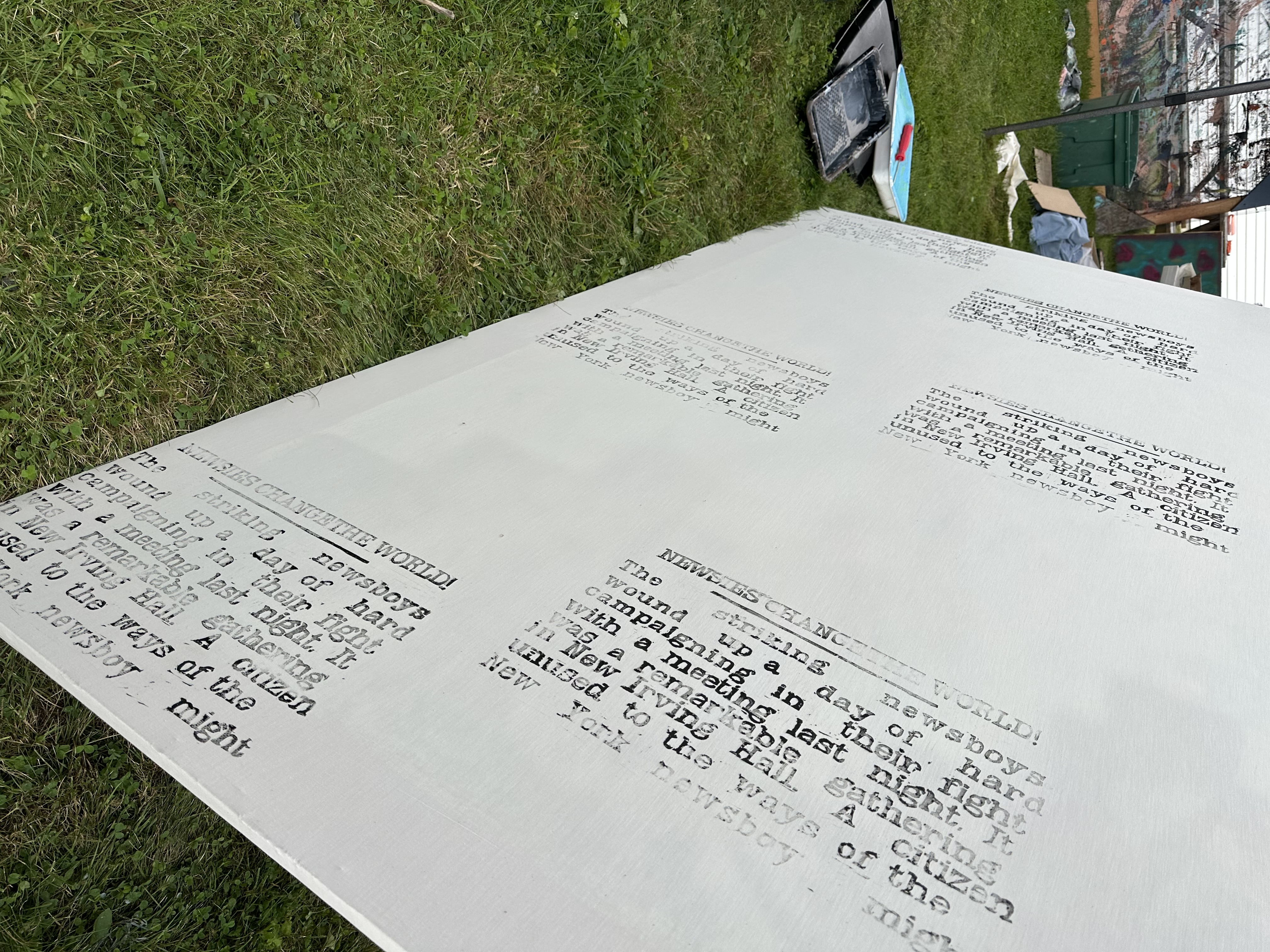 A big rectangular canvas laying on it's side in the grass with other set building stuff beyond it. The photo is turned at a weird angle so one can see the words that were printed. It is the first block over and over leaving a lot of white space under them and some in between. Look at the previous image for the full text - it's a lot!