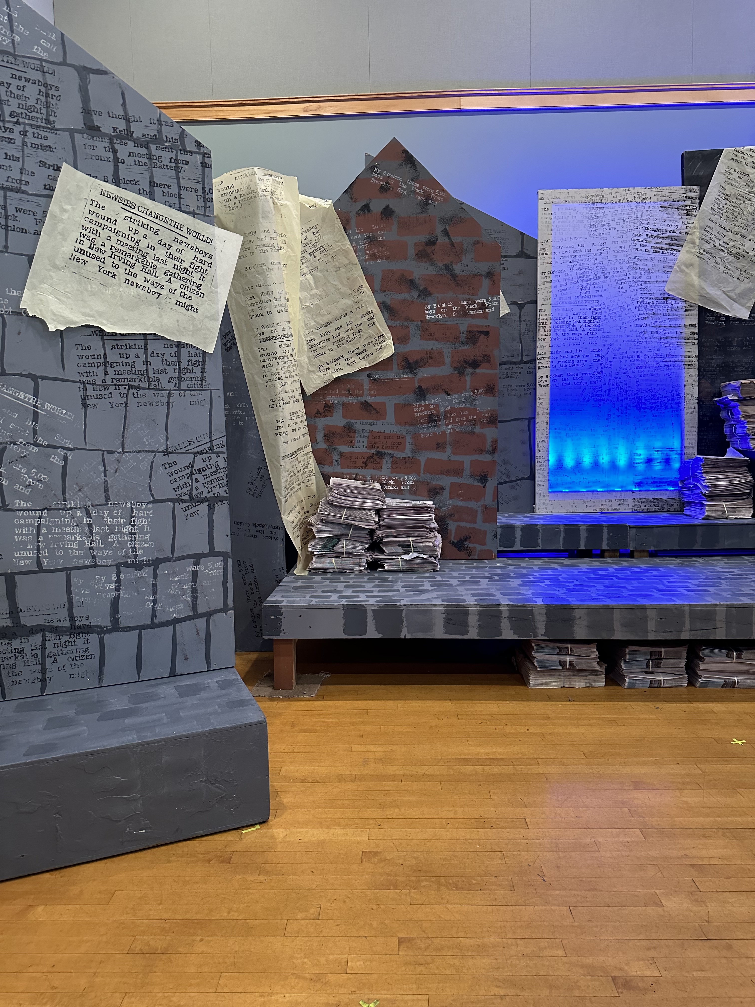 A part of the set for Newsies - panels with different angles are lined up. They are painted in various ways to appear as bricks and blocks of buildings. There is paper strewn across with the printing from the blocks on it and there's printing from parts of the blocks all over the panels in slightly different colors. The big canvas is to the right with blue lights behind. There are actual newspapers piled up around.