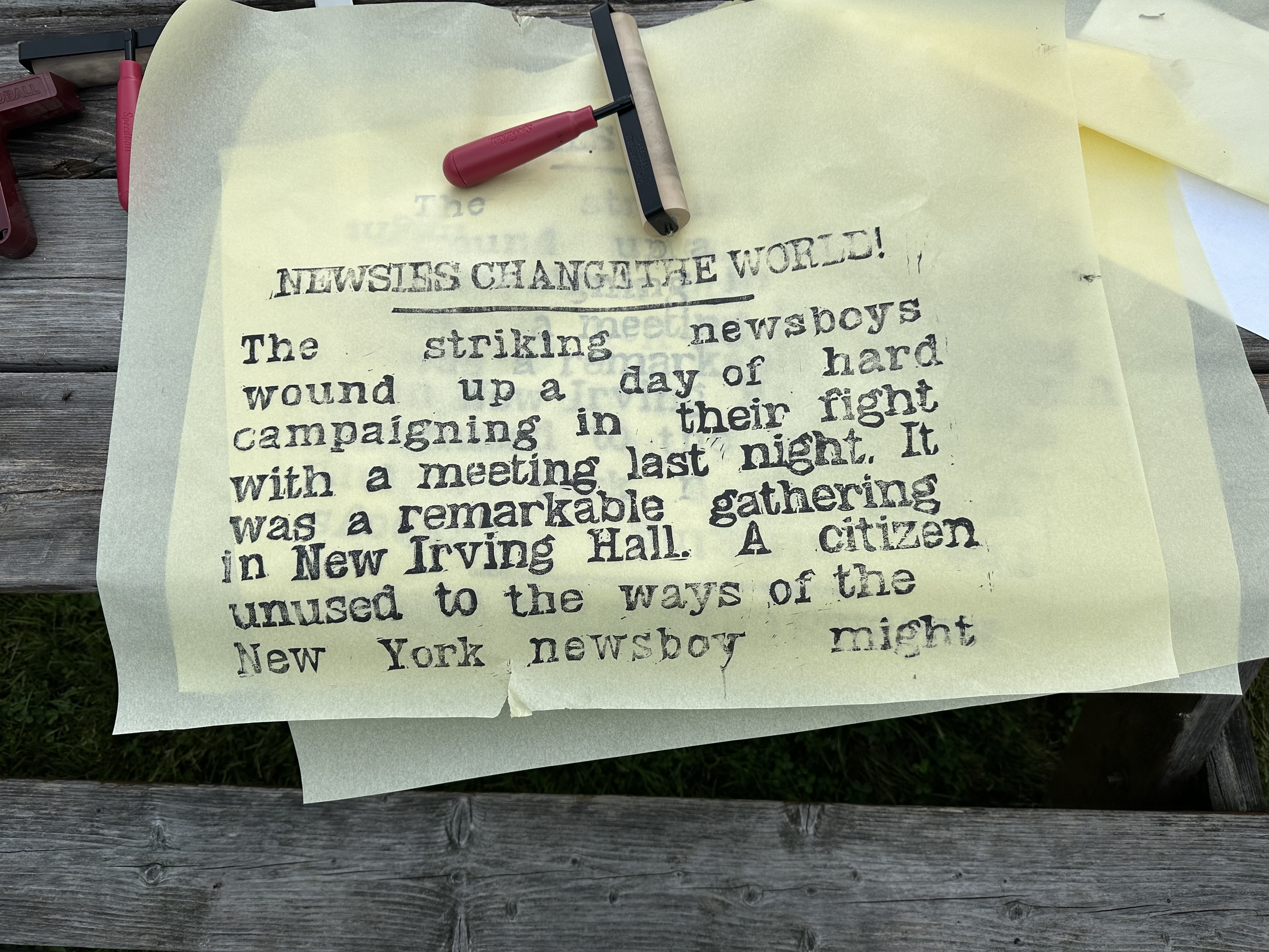 A test print on thin, beige paper of the first block. It's sitting on top of other pages on a wooden bench with a ink breyer on a blank space above. It says "NEWSIES CHANGE THE WORLD!" with a line under at the top. Under it says "The striking newsboys wound up a day of hard campaigning in their fight with a meeting last night. It was a remarkable gathering in New Irving Hall. A citizen unused to the ways of the New York newsboy might"