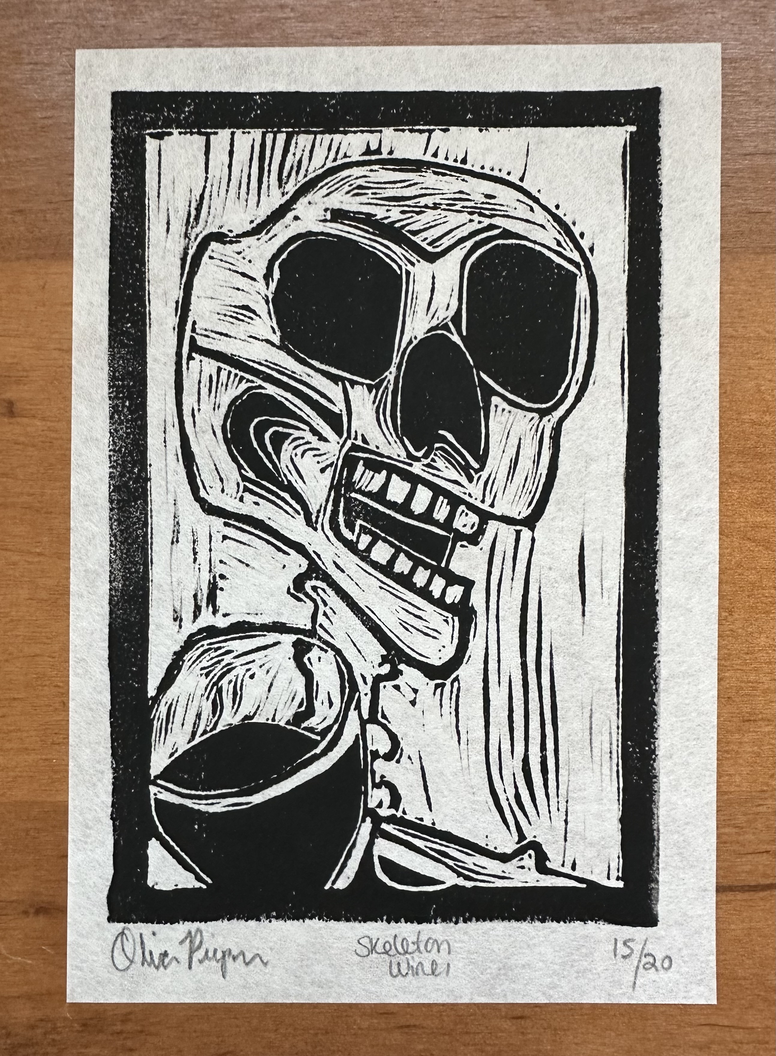 A block print of a skeleton from the shoulders up, though their left shoulder and neck are mostly covered by what looks like a glass of wine. Underneath the print is a black blob on the left (my signature blacked out), "Skeleton Wine I" in the center, and 15/20 to the right,