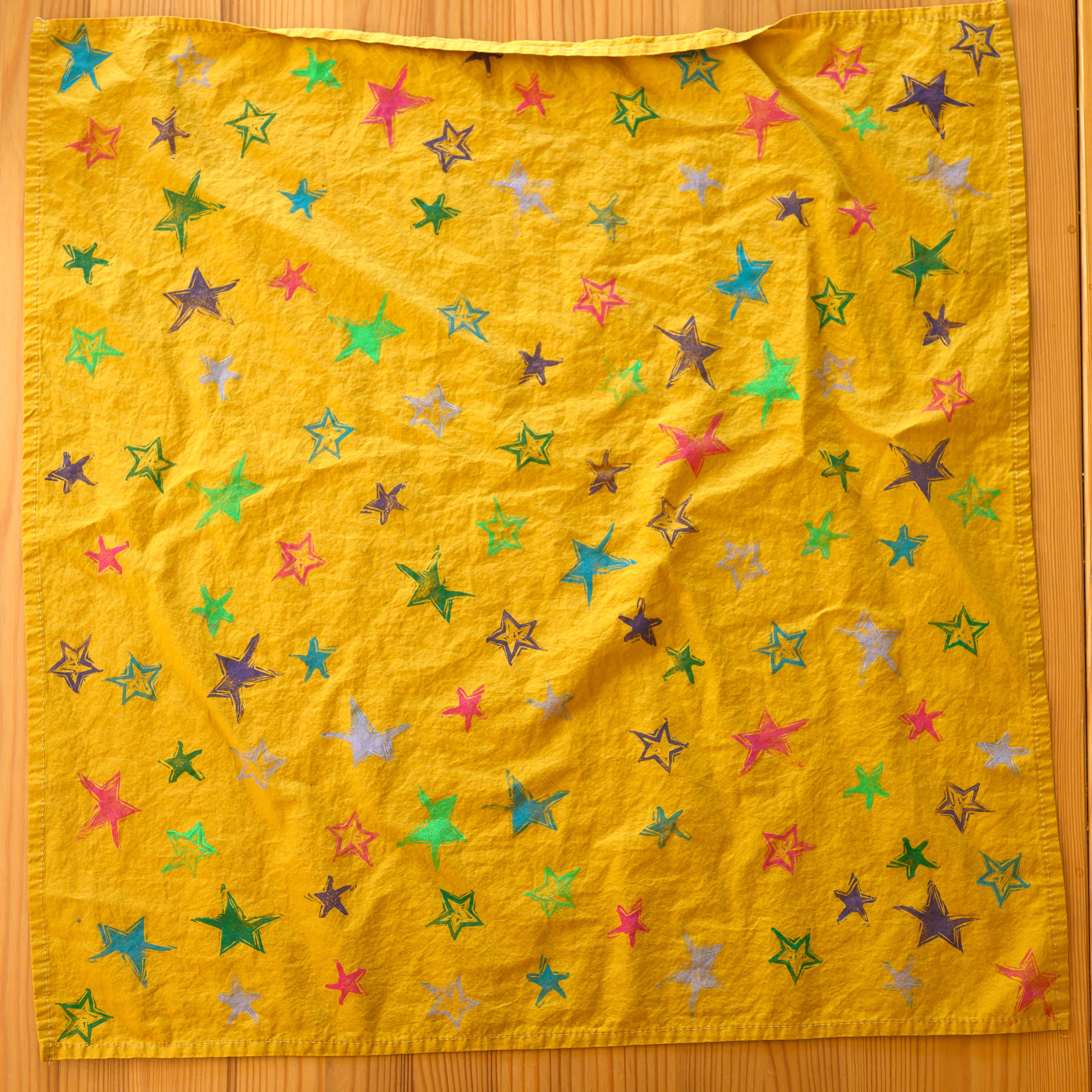 A mustard yellow bandana from above. It is covered in stars of various colors and styles.
