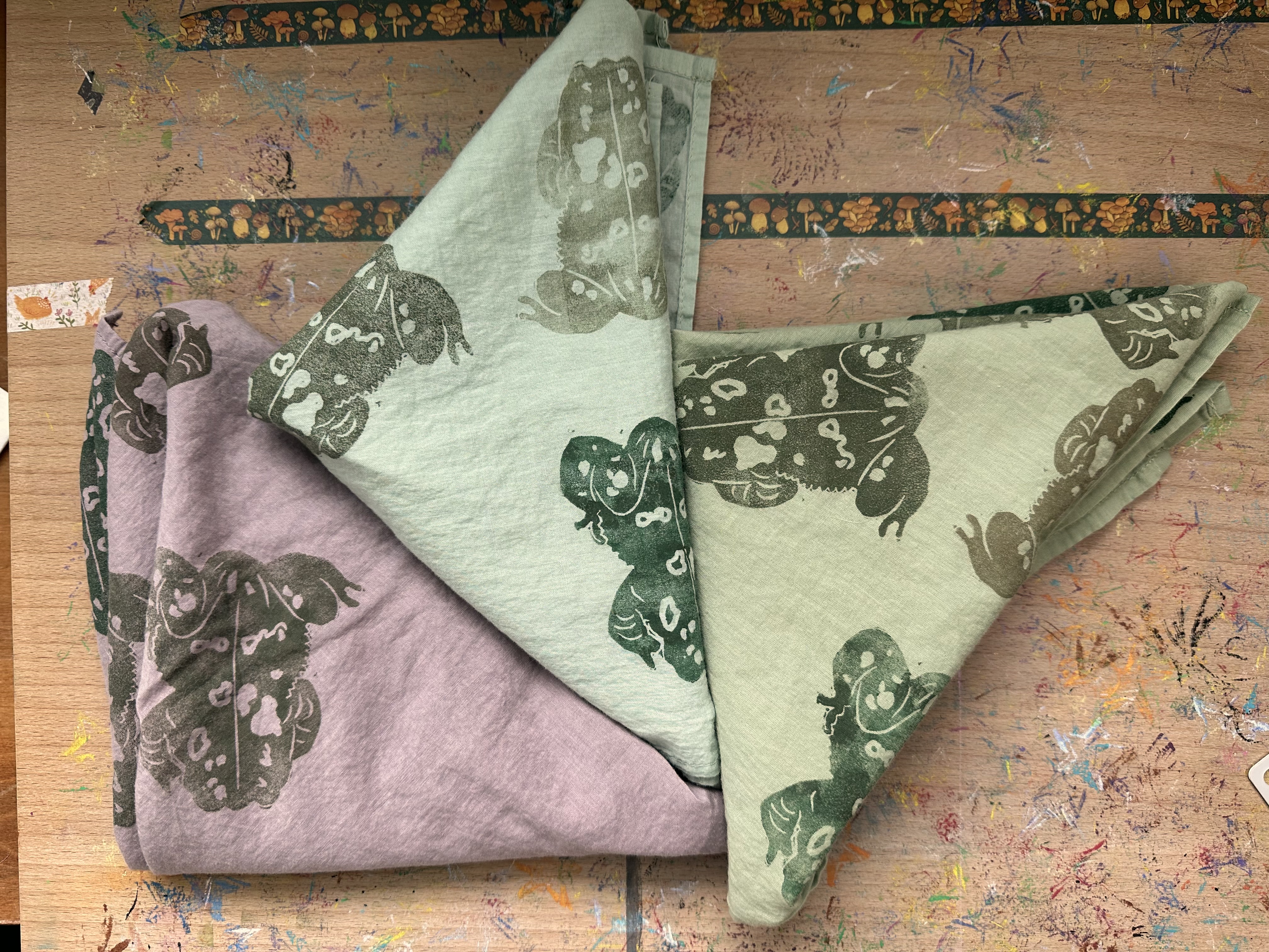 Three bandanas folded into triangles from above. They all have toads of various greens/browns on them. The bandanas are dusky pink, light green blue, and light green.