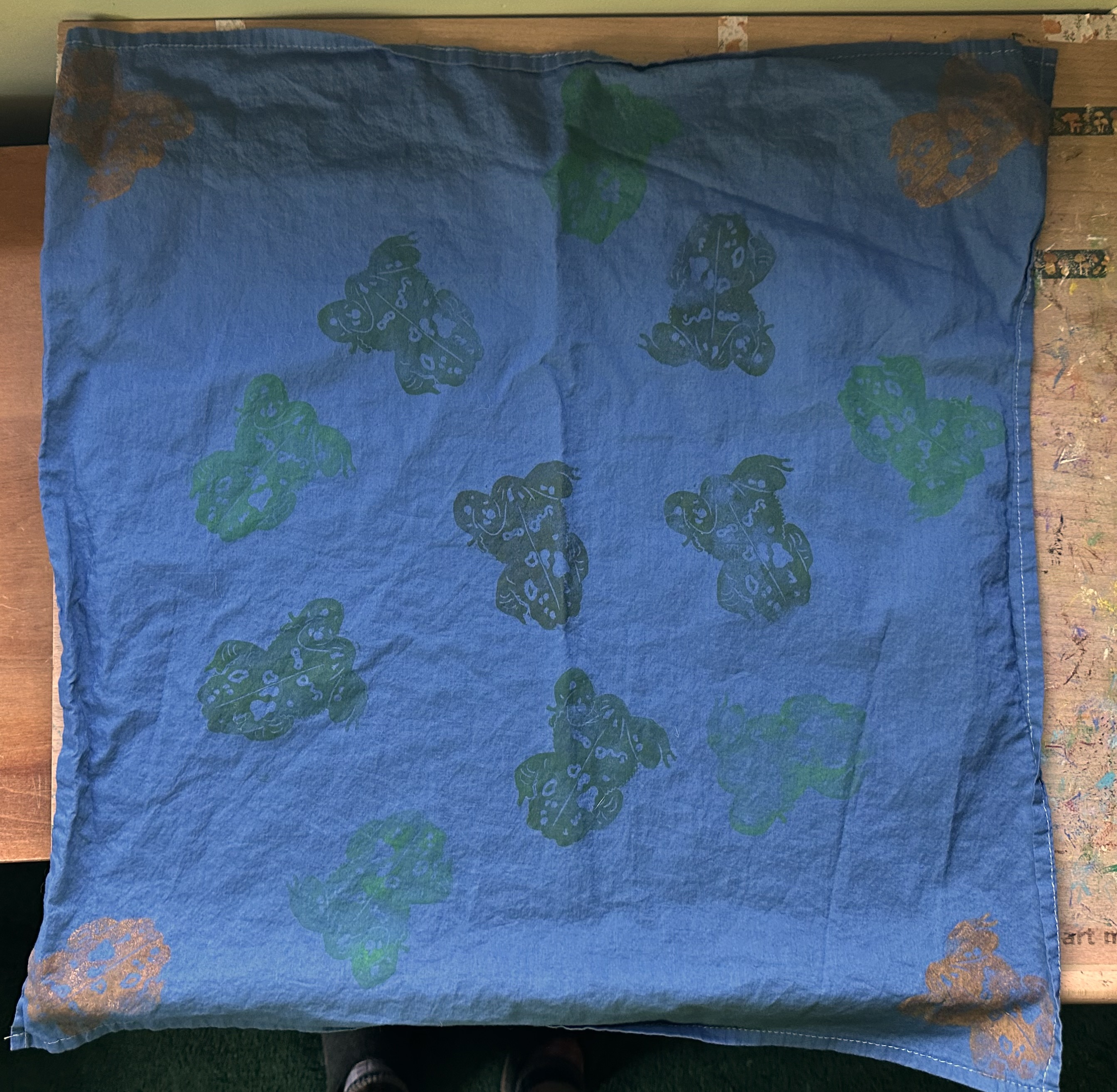A dusky blue bandana from above with toads on it. Tan/brown ones in the corners and in the center is an assortment of green/brown ones.