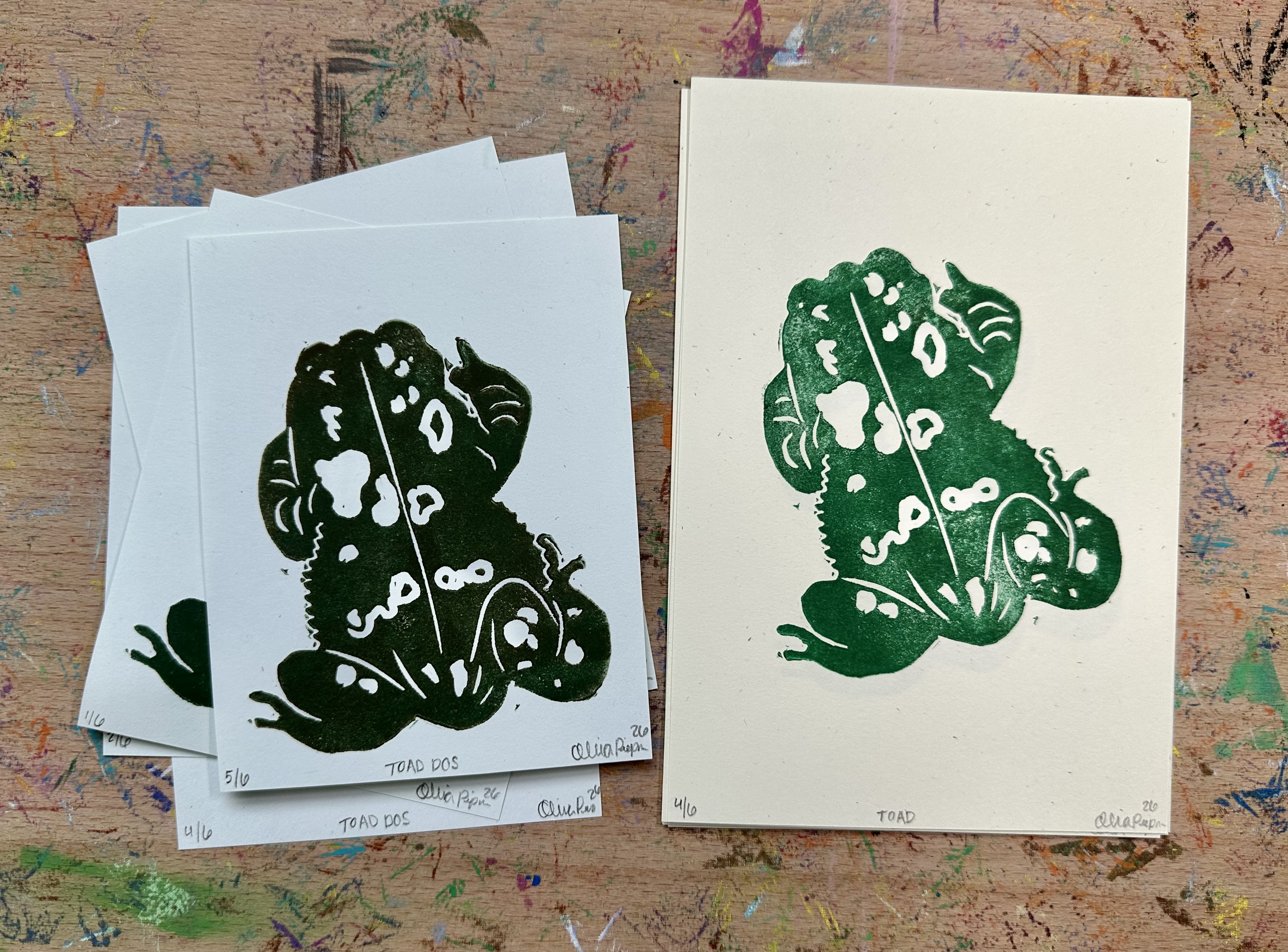 Two stacks of block prints of the same toad. The stack on the left is on blue-ish white paper and the toad is a very dark green/brown. The stack on the right is on bigger slightly yellow/green white paper and the toad is greener.